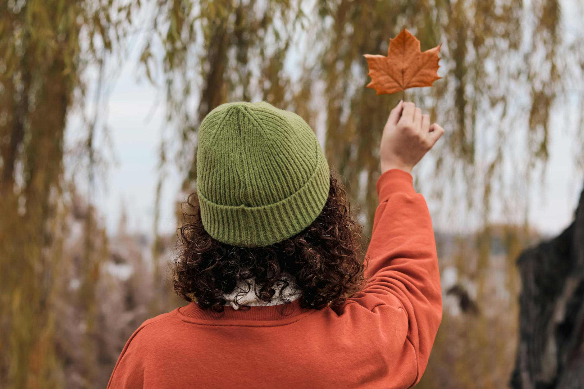 A woman in a green hat holds up a maple leaf, embracing the fall season.