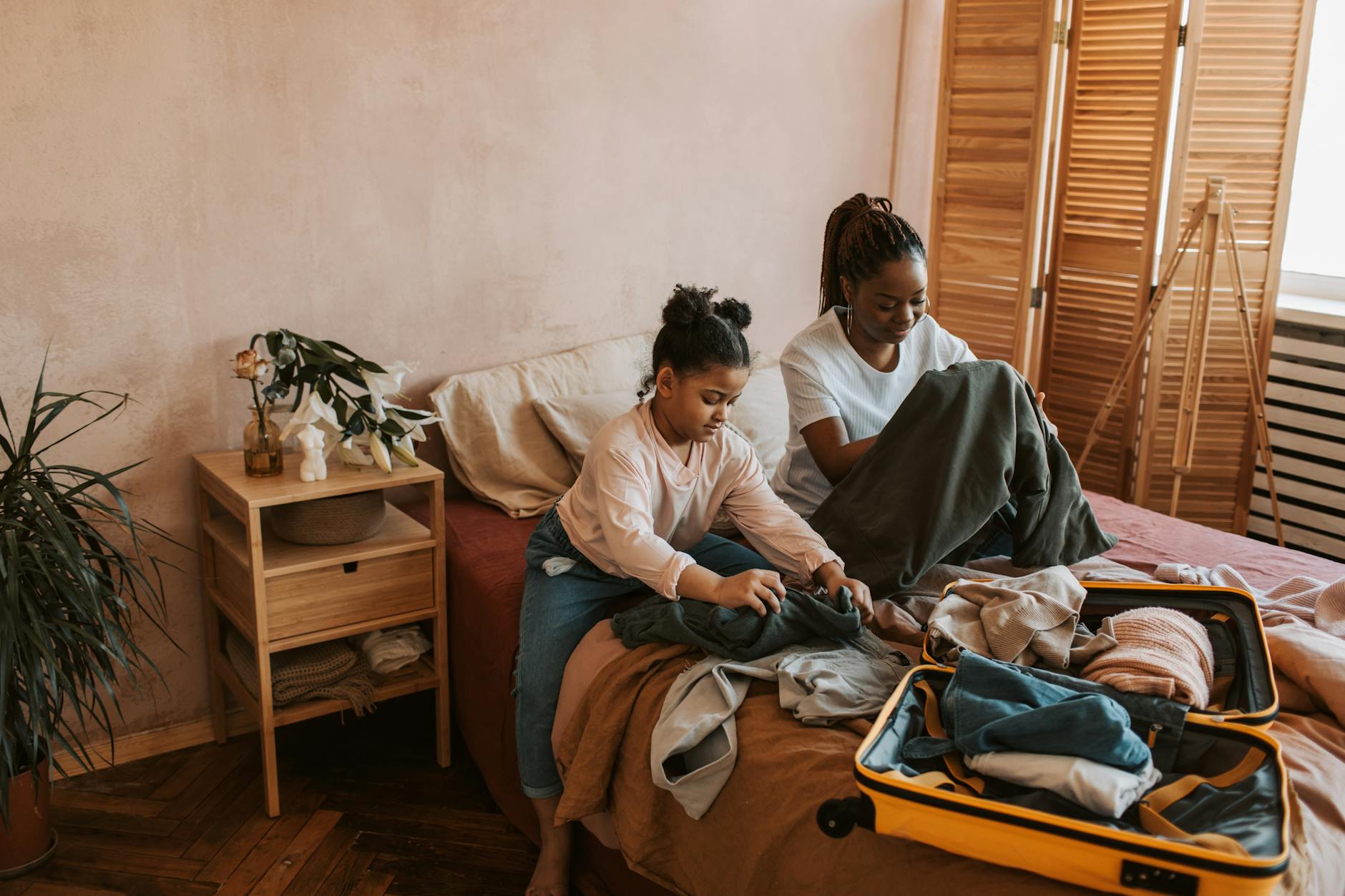 A mother and daughter fold clothes while packing a suitcase on a bed indoors.
