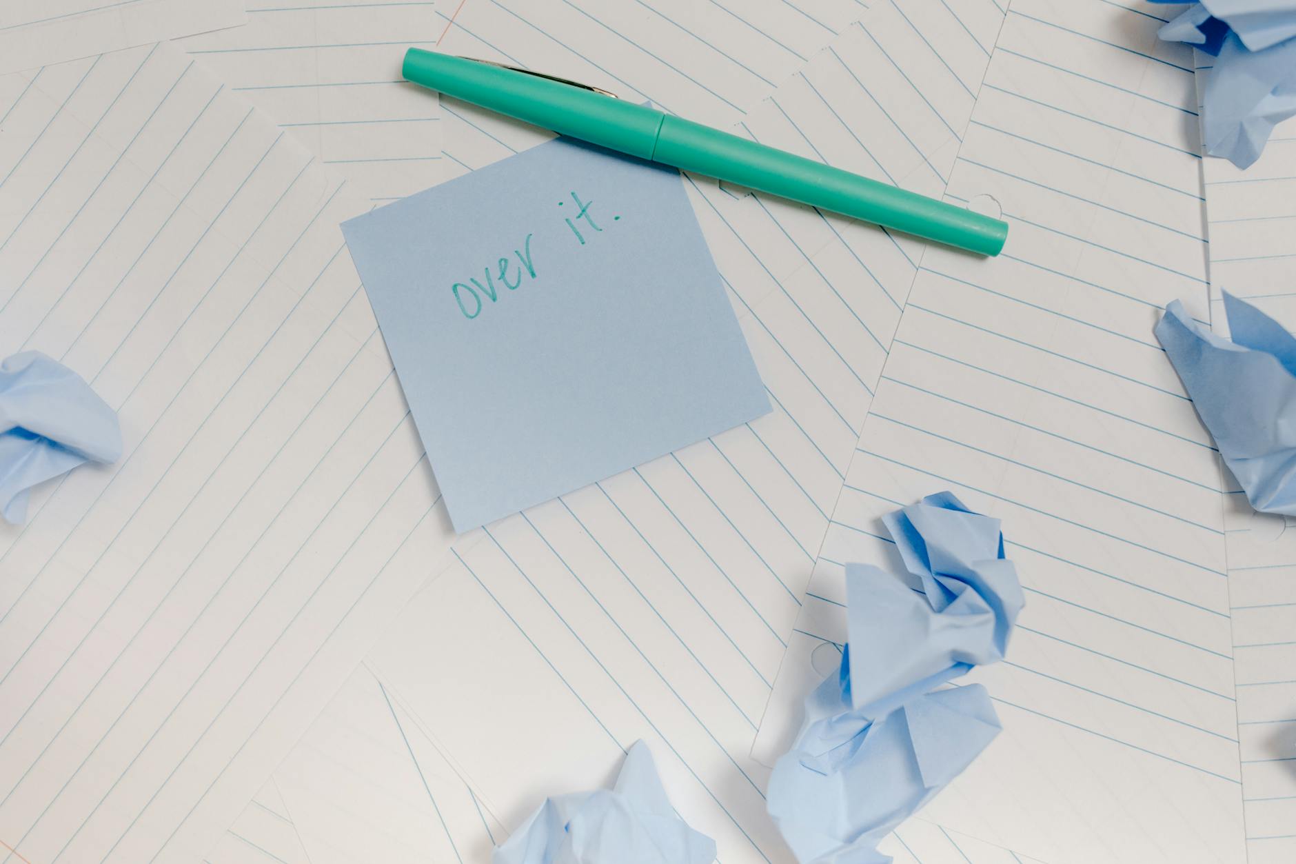 Image of a blue sticky note with 'over it' text, crumpled papers, and a turquoise pen on lined paper.
