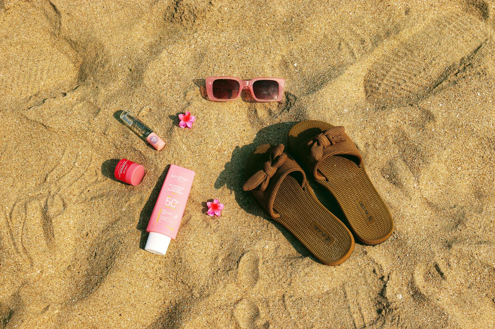 A flat lay of summer beach essentials including sandals, pink sunglasses, and sunscreen on sandy beach.
