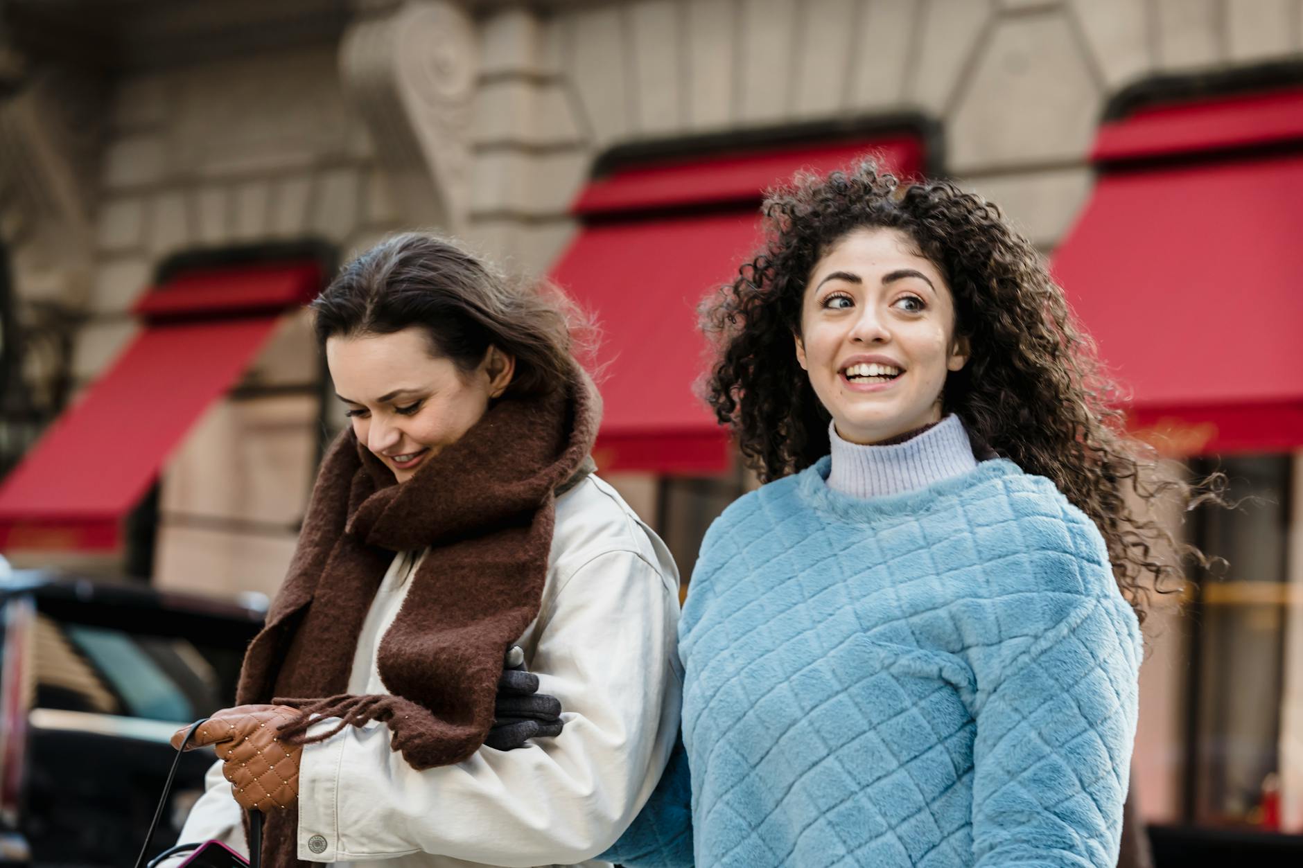 Cheerful trendy young multiethnic female millennials in stylish warm clothes smiling while strolling on city street near aged building