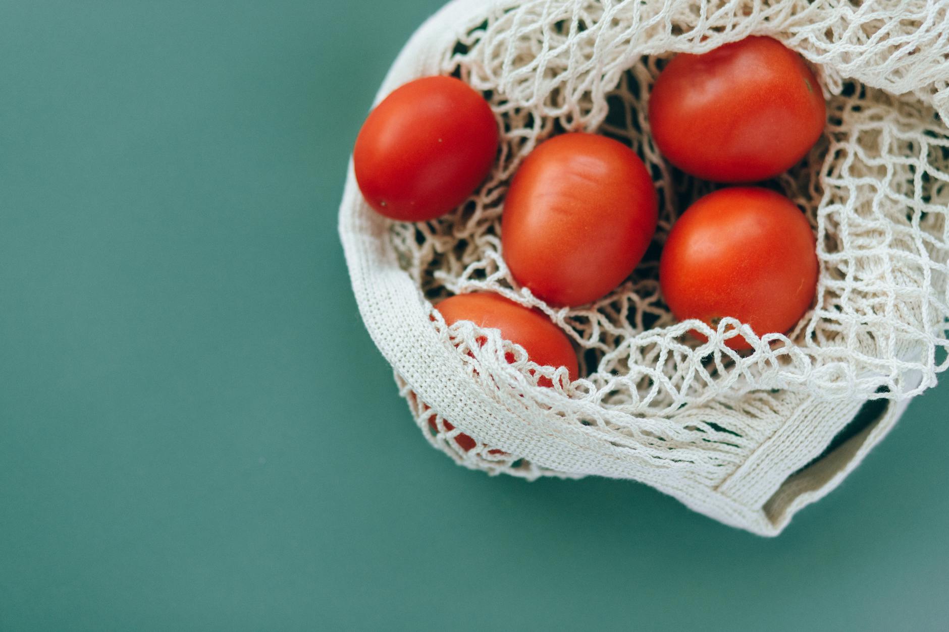 Aerial view of ripe tomatoes in a reusable mesh bag on a green surface, emphasizing freshness and eco-friendliness.